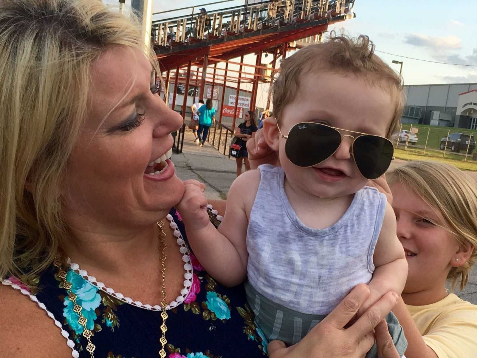 mother smiles while holding adopted infant with aviator sunglasses on and sister laughing