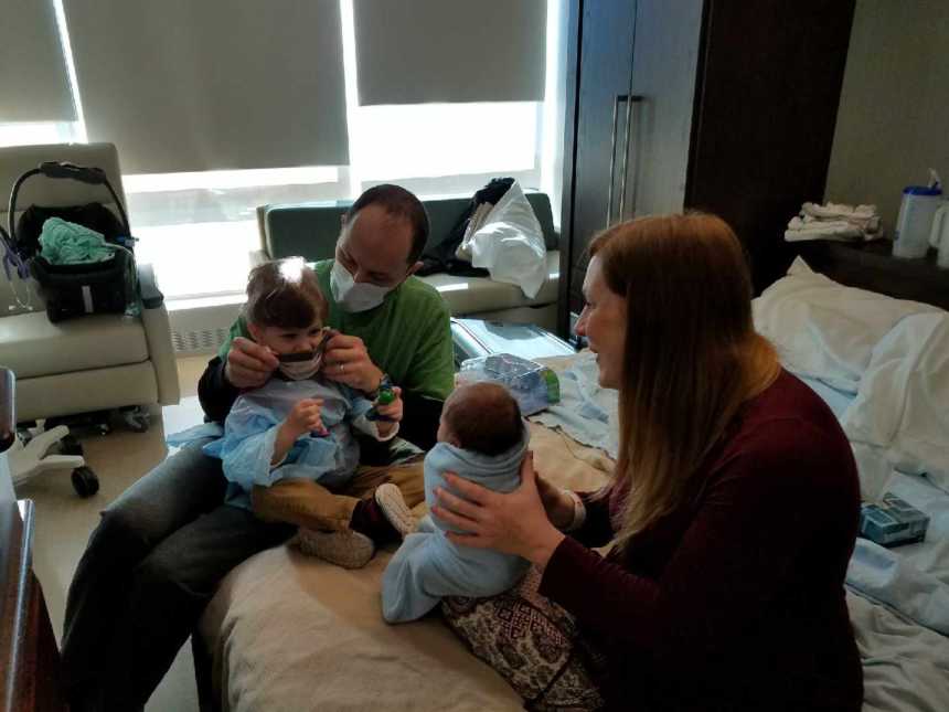 man and woman hold a toddler and newborn in their laps in a hospital room