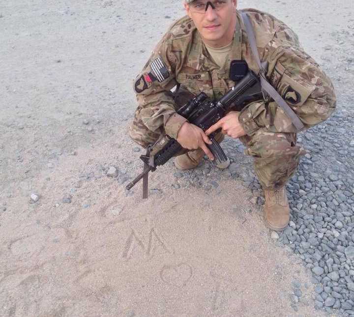 soldier kneels in sand where he drew an M, a heart ,and a T with his gun