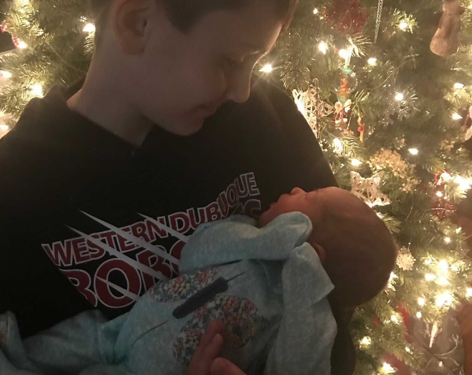 brother holds adopted baby sister in his arms in front of Christmas tree