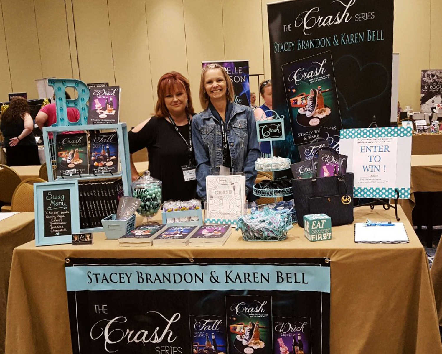 two women smiling behind a table with books on it and a poster with cover of book on it