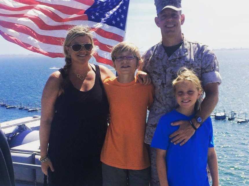 Marine husband and dad smiles with wife, son, and daughter with American flag behind them and ocean in background