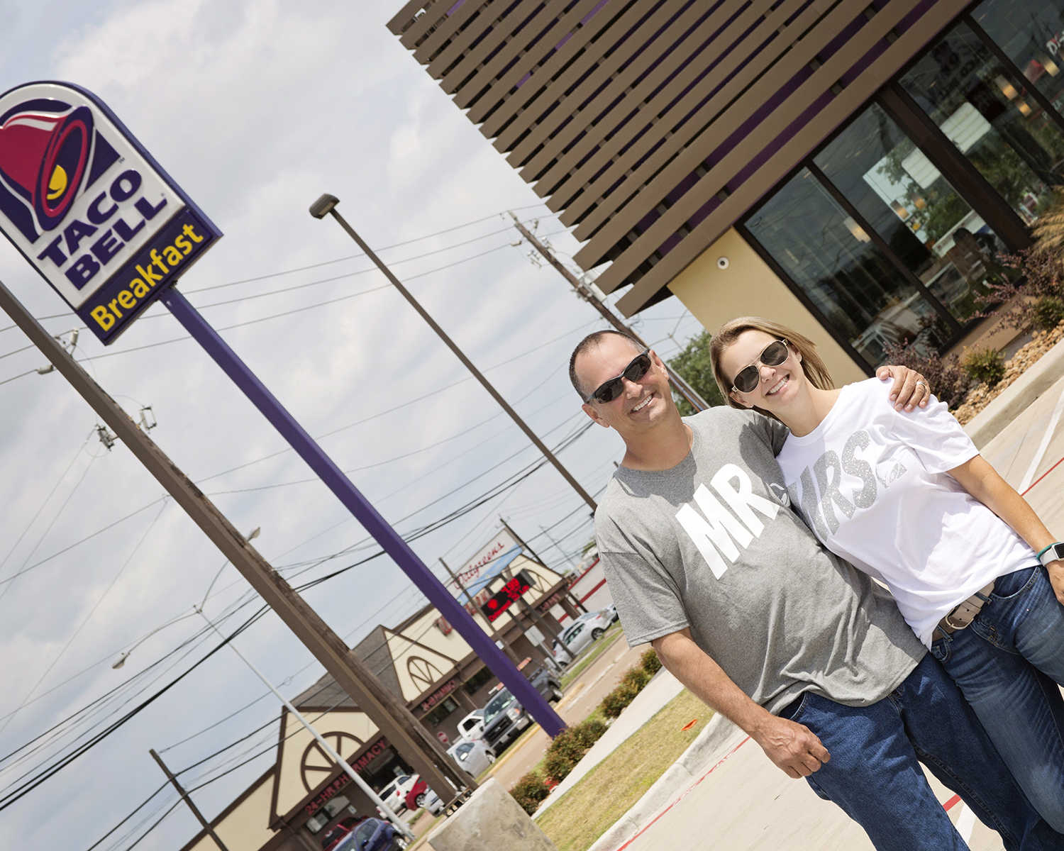 husband and wife smiling in front of taco bell sign wearing shirts that read, "Mr." and "Mrs."