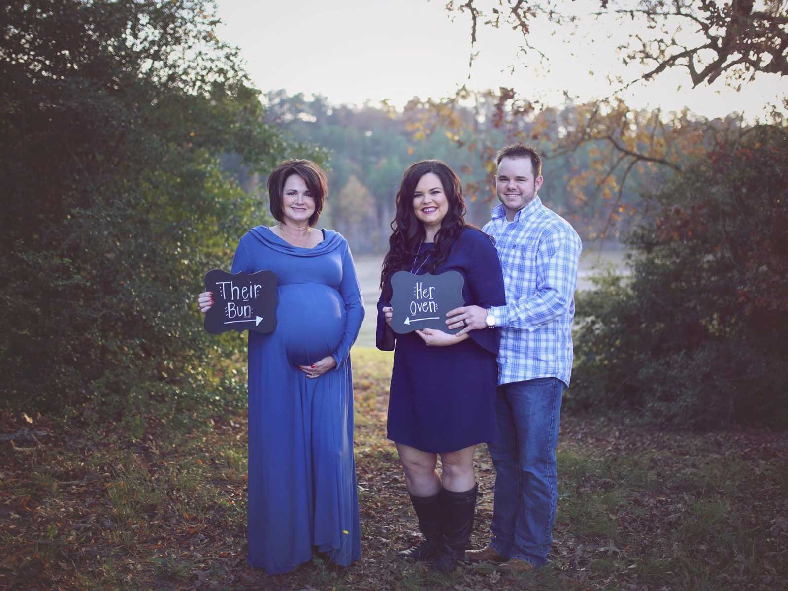 Pregnant woman stands in a field holding a sign saying, "their bun" and couple to the right holds a sign saying, "her oven"