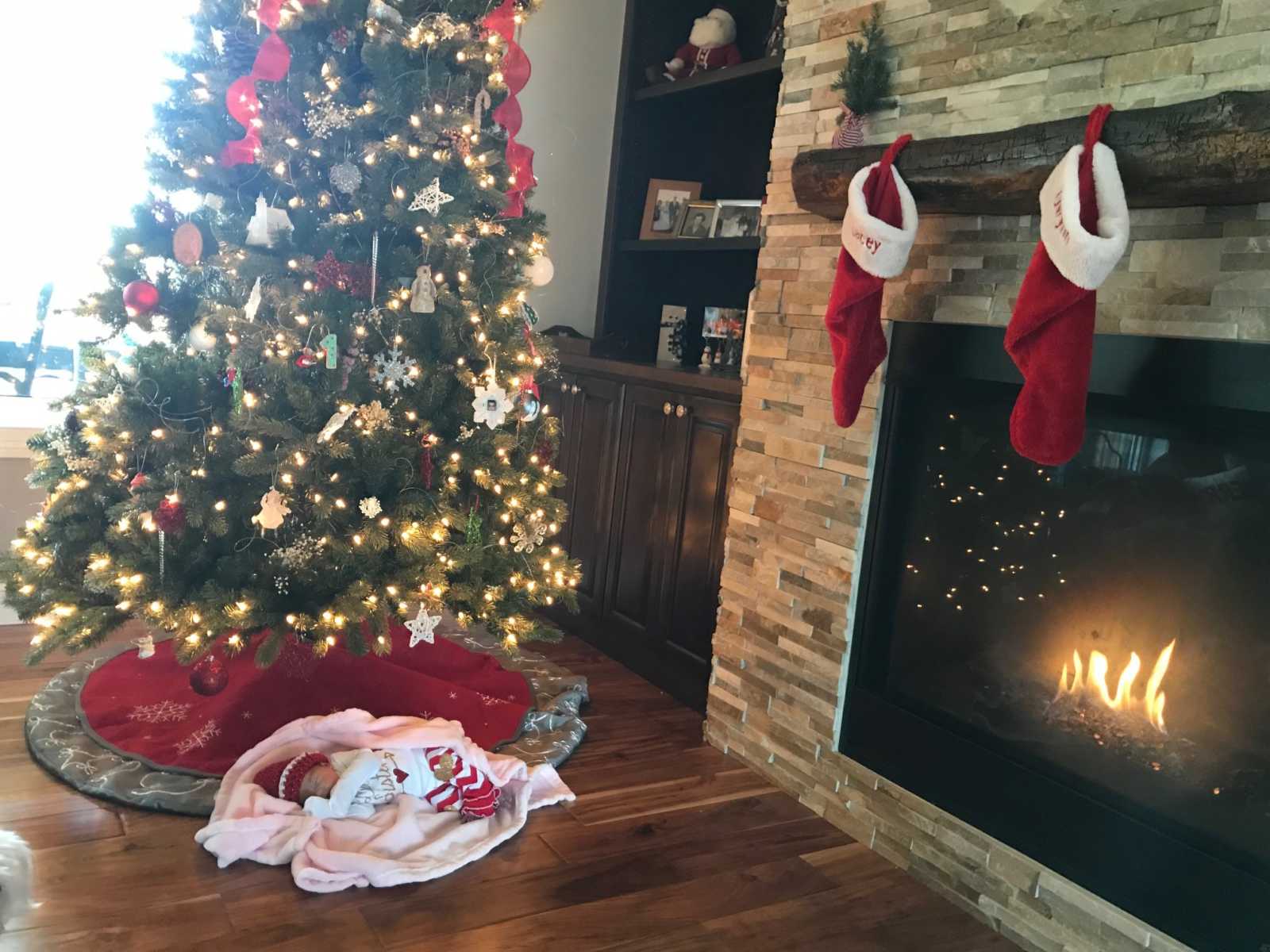 adopted newborn baby is wrapped in blankets under Christmas tree next to a burning fireplace and stockings