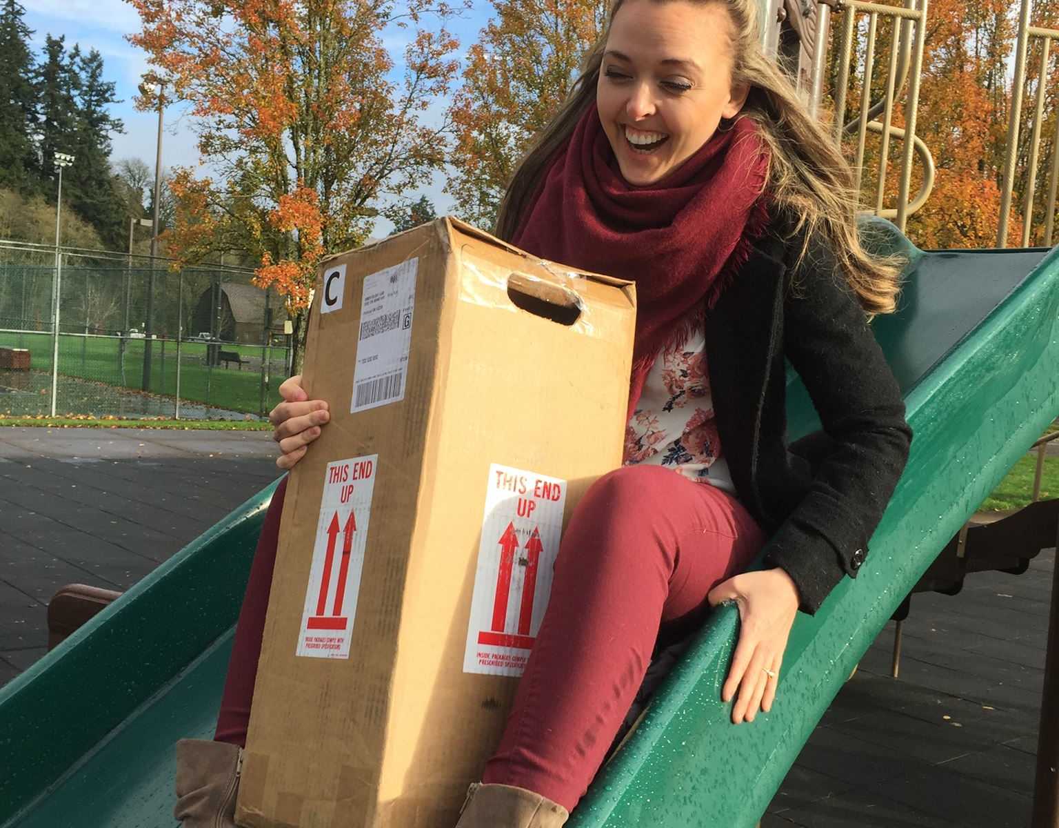 woman smiling and going down a slide at a playground while holding a box of embryos