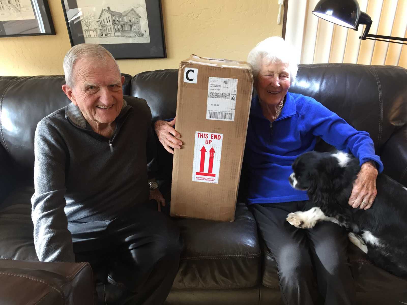 elderly man and woman sit on a couch with a dog and a box of embryos is sitting in between them