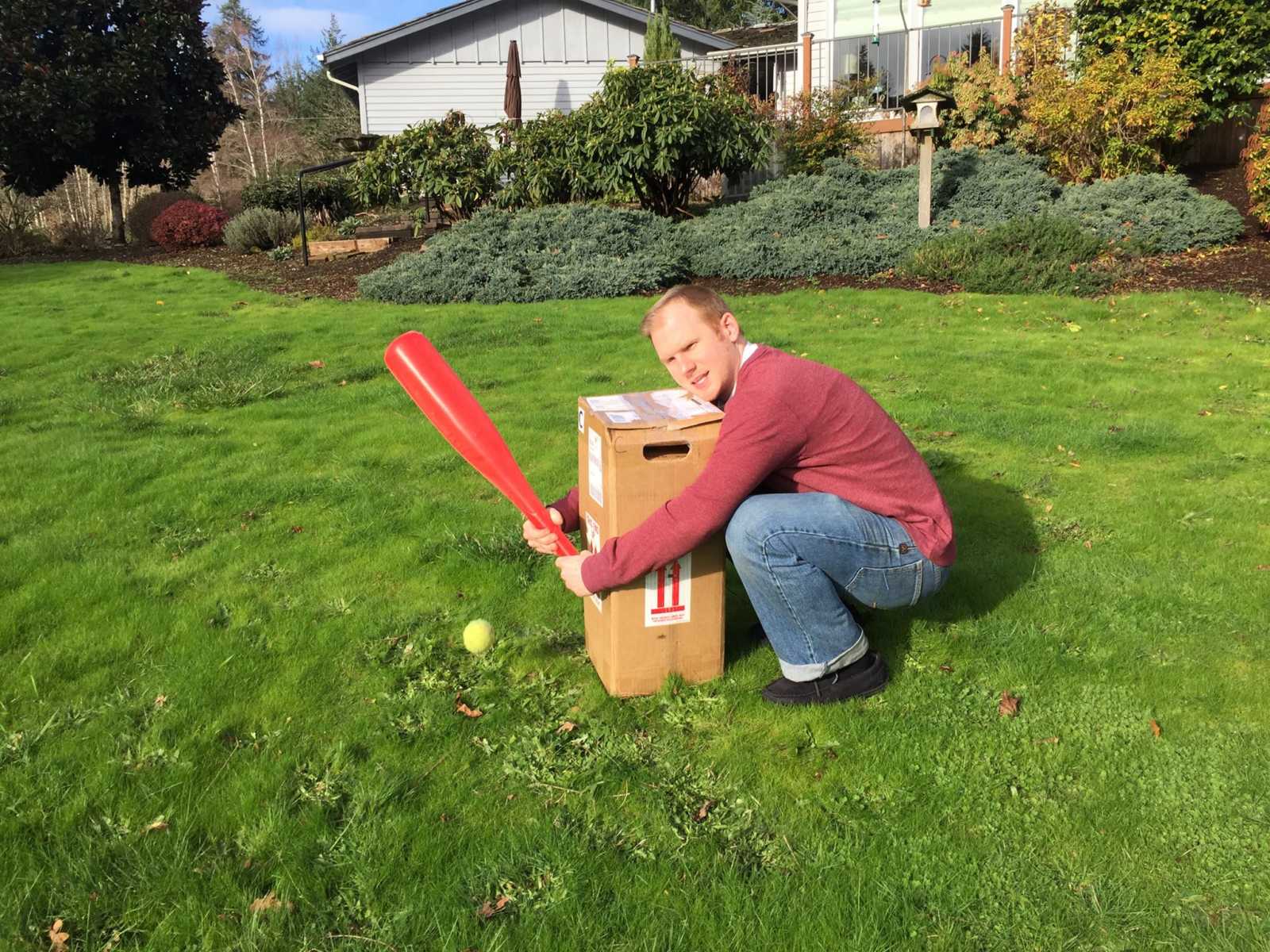 man crouches down behind a box with his arms around it and a plastic basball bat in his hands and ball on the ground