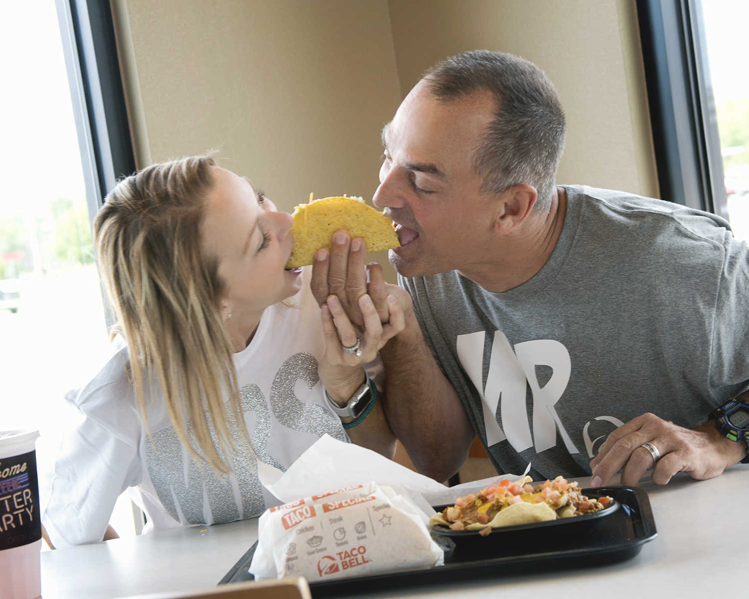 husband and wife take bite of opposite end of taco with a tray off food in front of them at taco bell