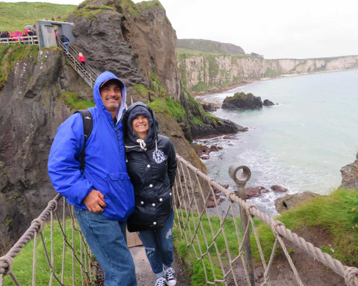husband and wife in raincoats on a roped bridge crossing over a body of water on a cliff