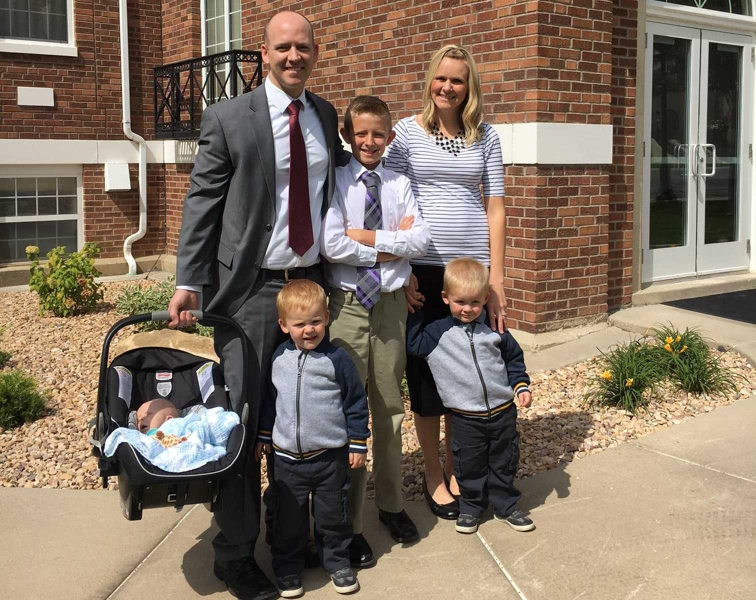 father stands next to older son and wife with twins in front of them while holding baby in carseat