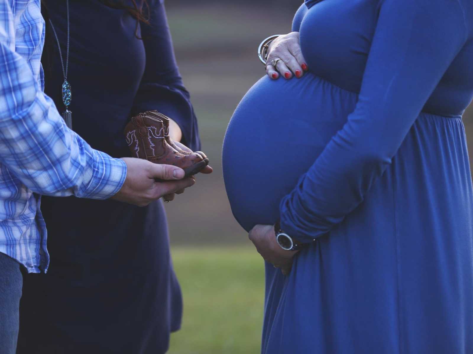 pregnant woman holds her stomach while man and woman hold baby cowboy boots in their hands