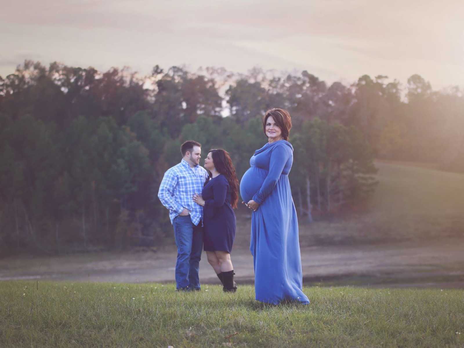 Pregnant woman stands in the forefront holding her stomach while man and woman are arm and arm in the background