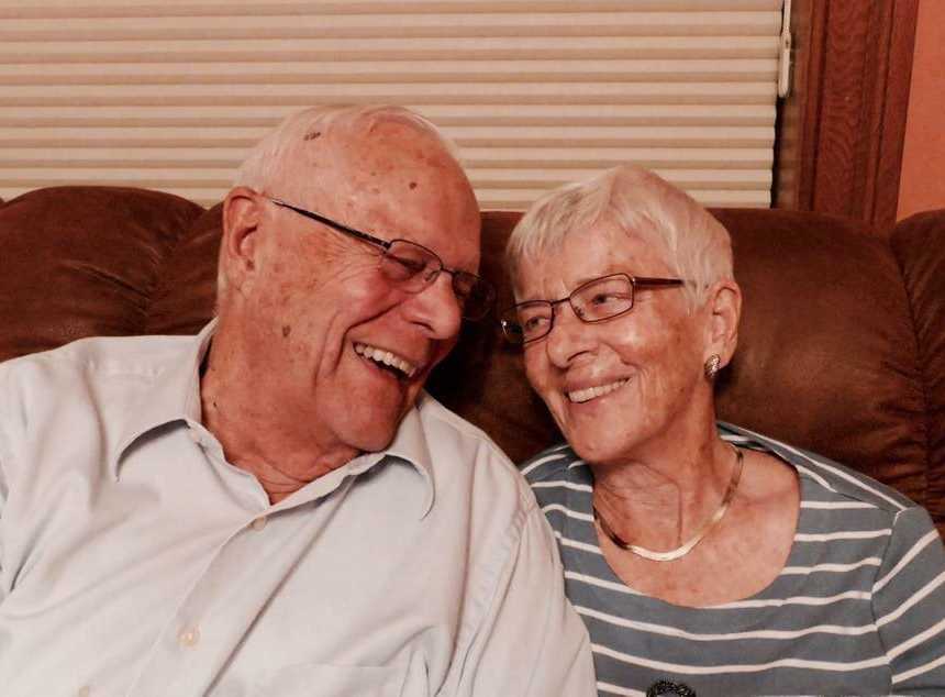 Woman with dementia sits on couch smiling at husband sitting next to her