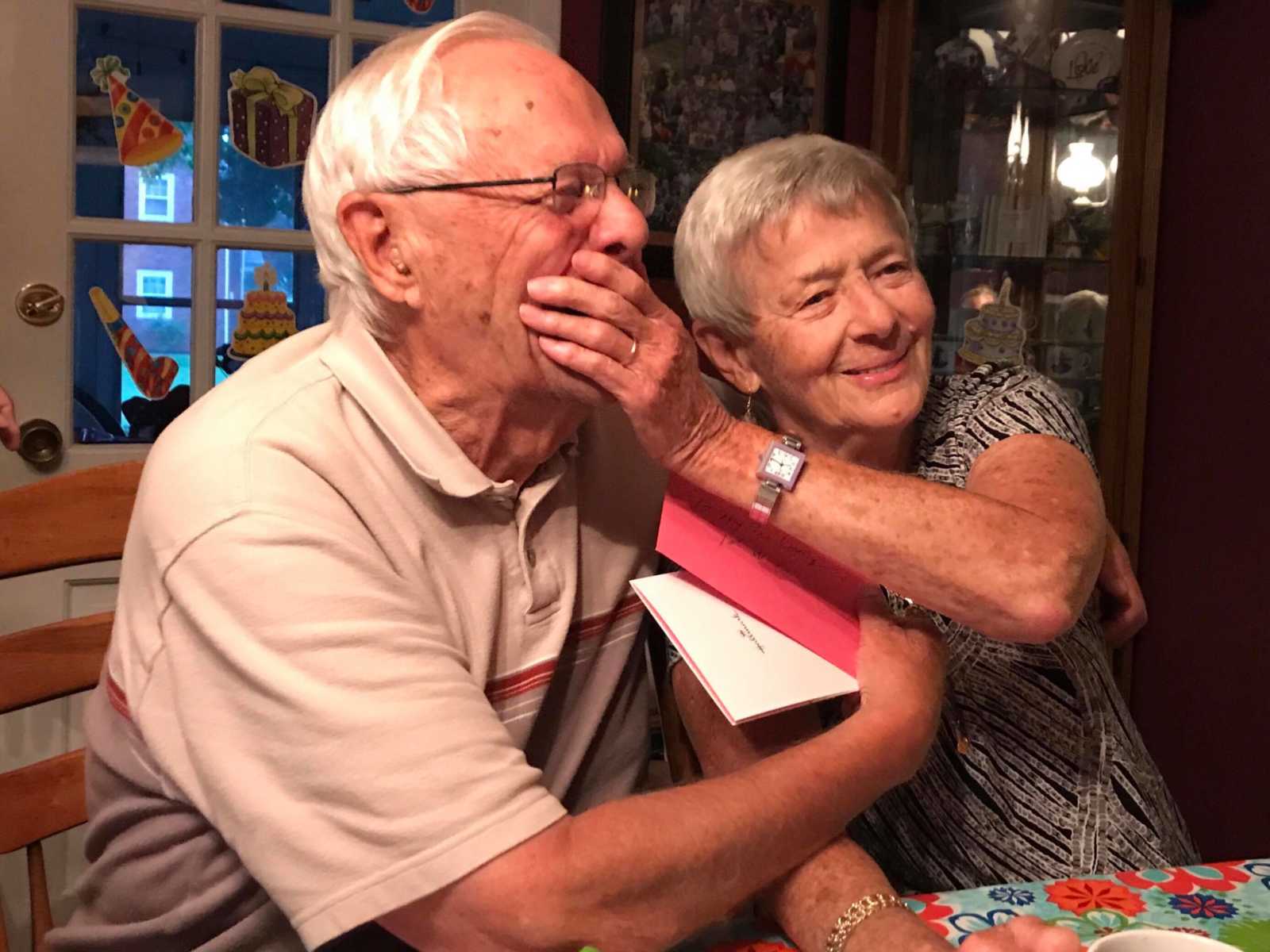 Elderly woman with dementia smiles while placing hand on the mouth of husband laughing
