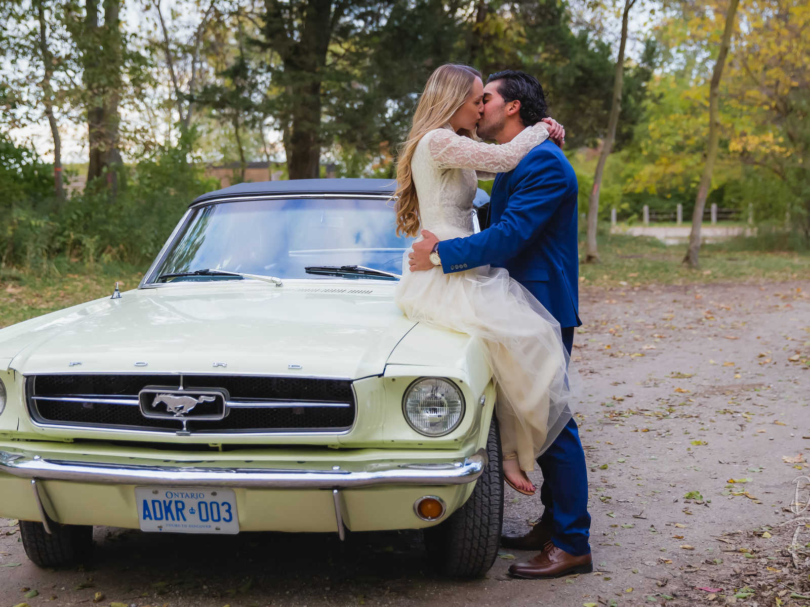 woman sits on an old yellow mustang while fiancee stands kissing her