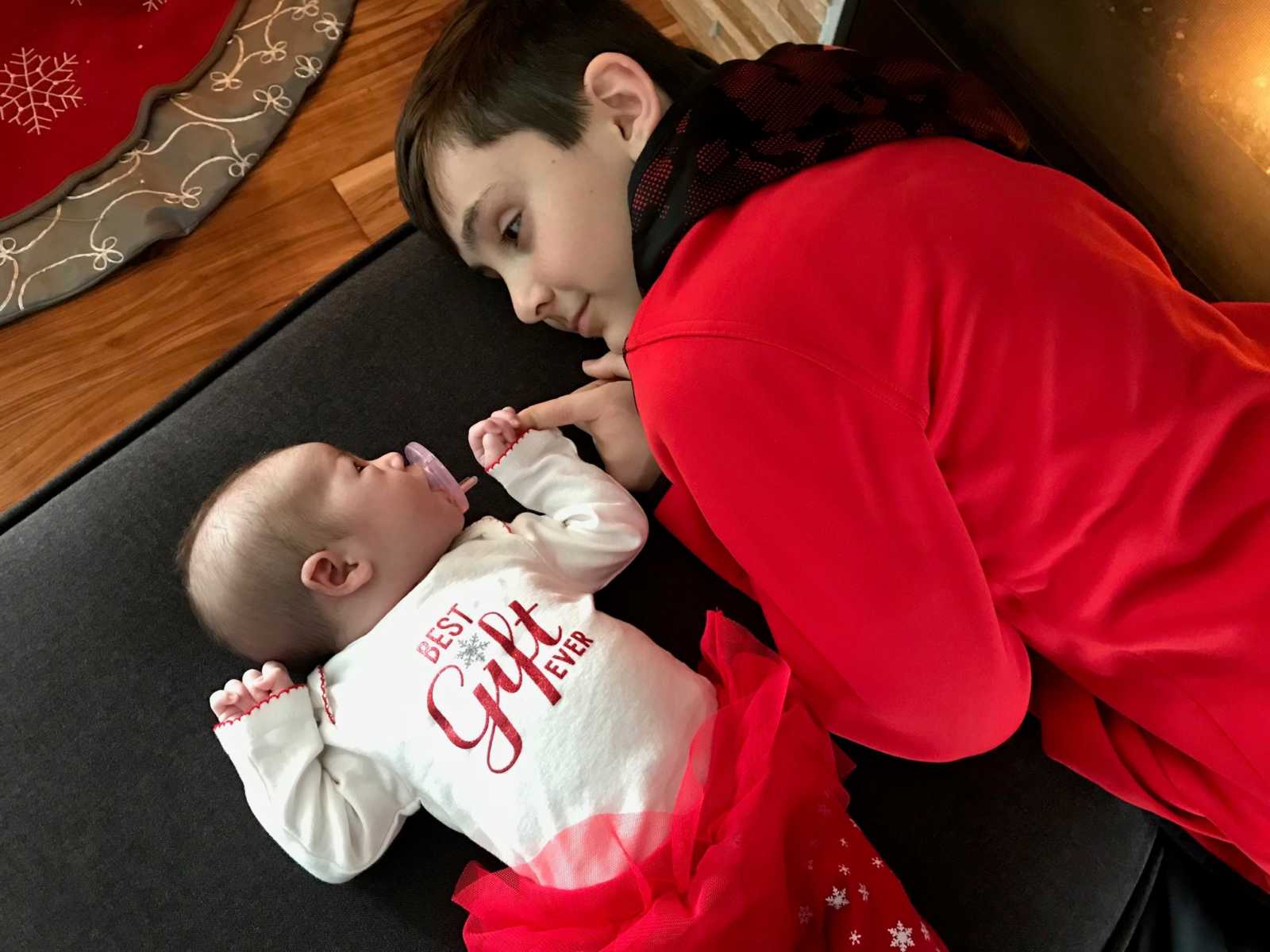 brother lays on the floor and holds hands with adopted baby sister with shirt that says, "best gift ever"
