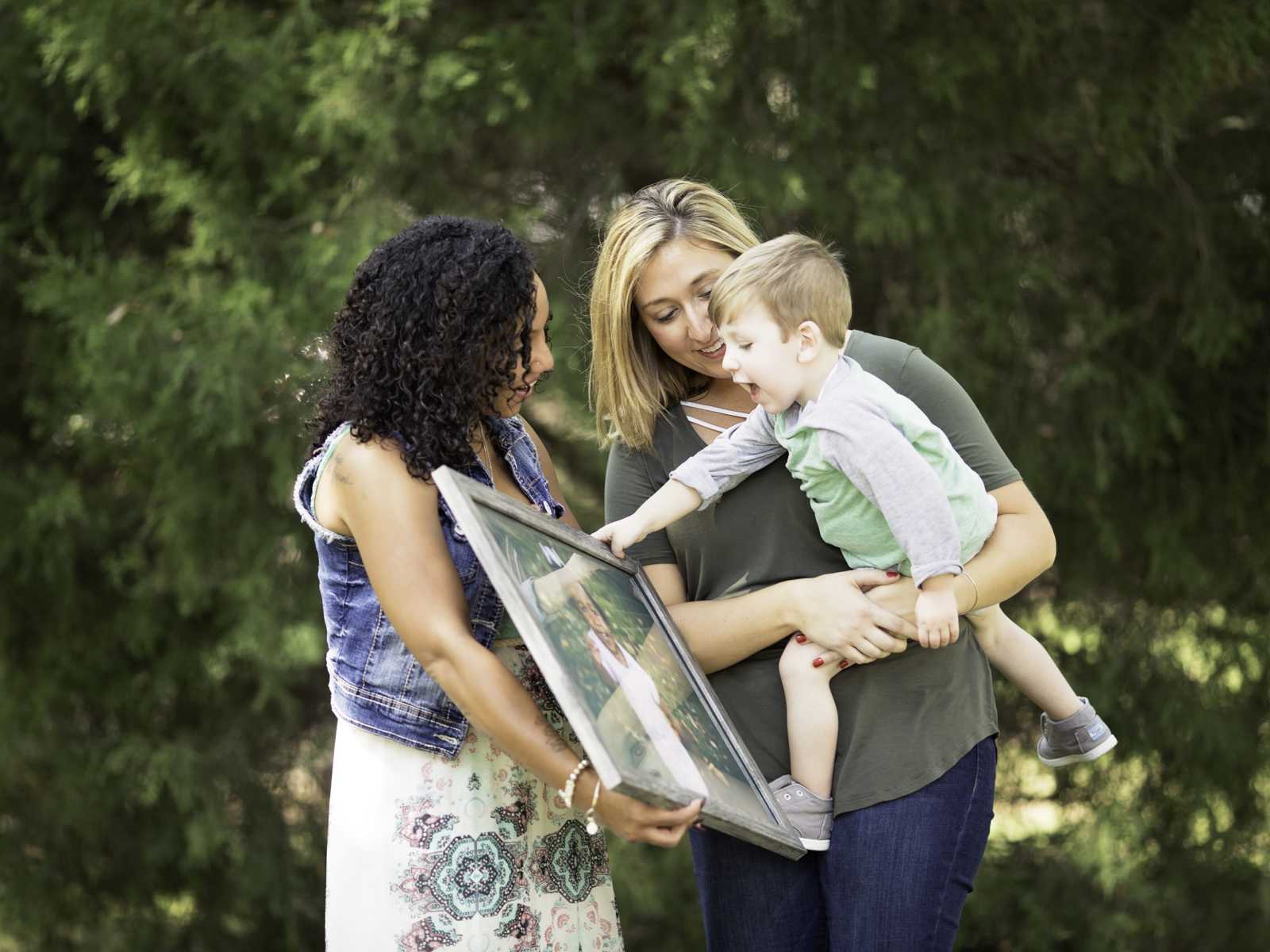Woman holds picture frame of deceased daughter and little boy in mom's arms points at it