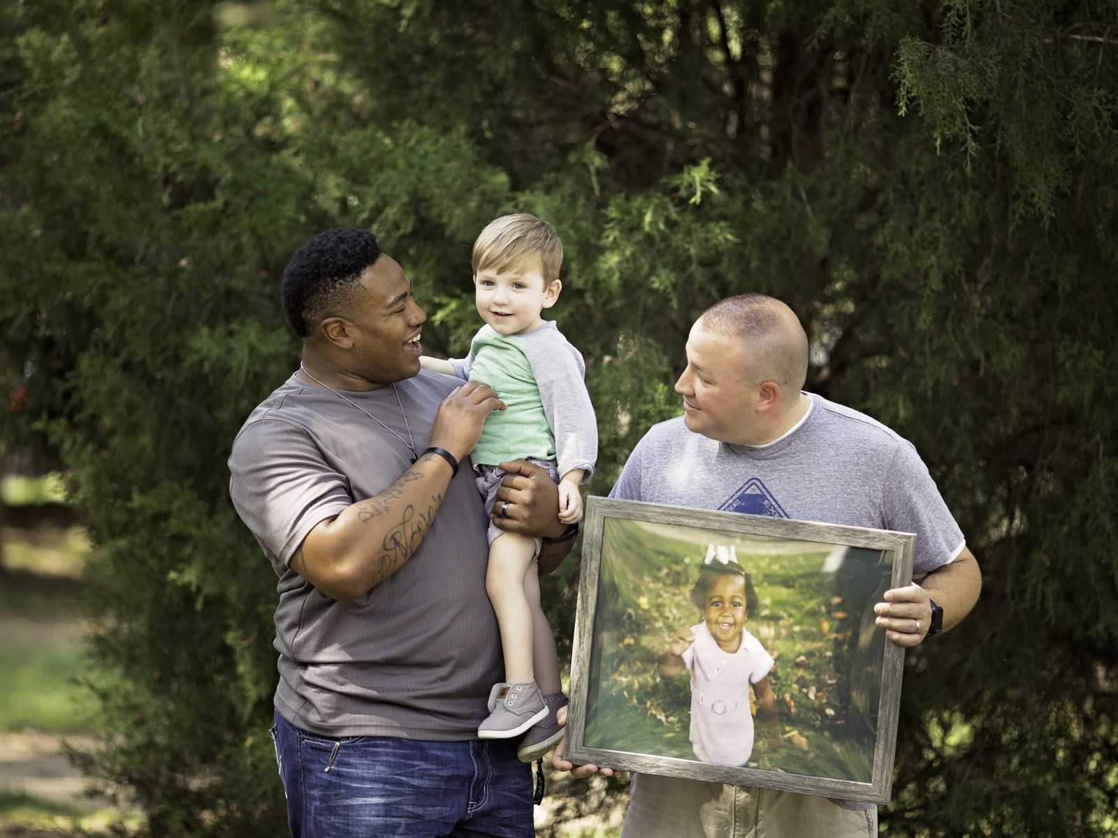 Man holds little boy whose father stands beside them holding picture frame of deceased little girl