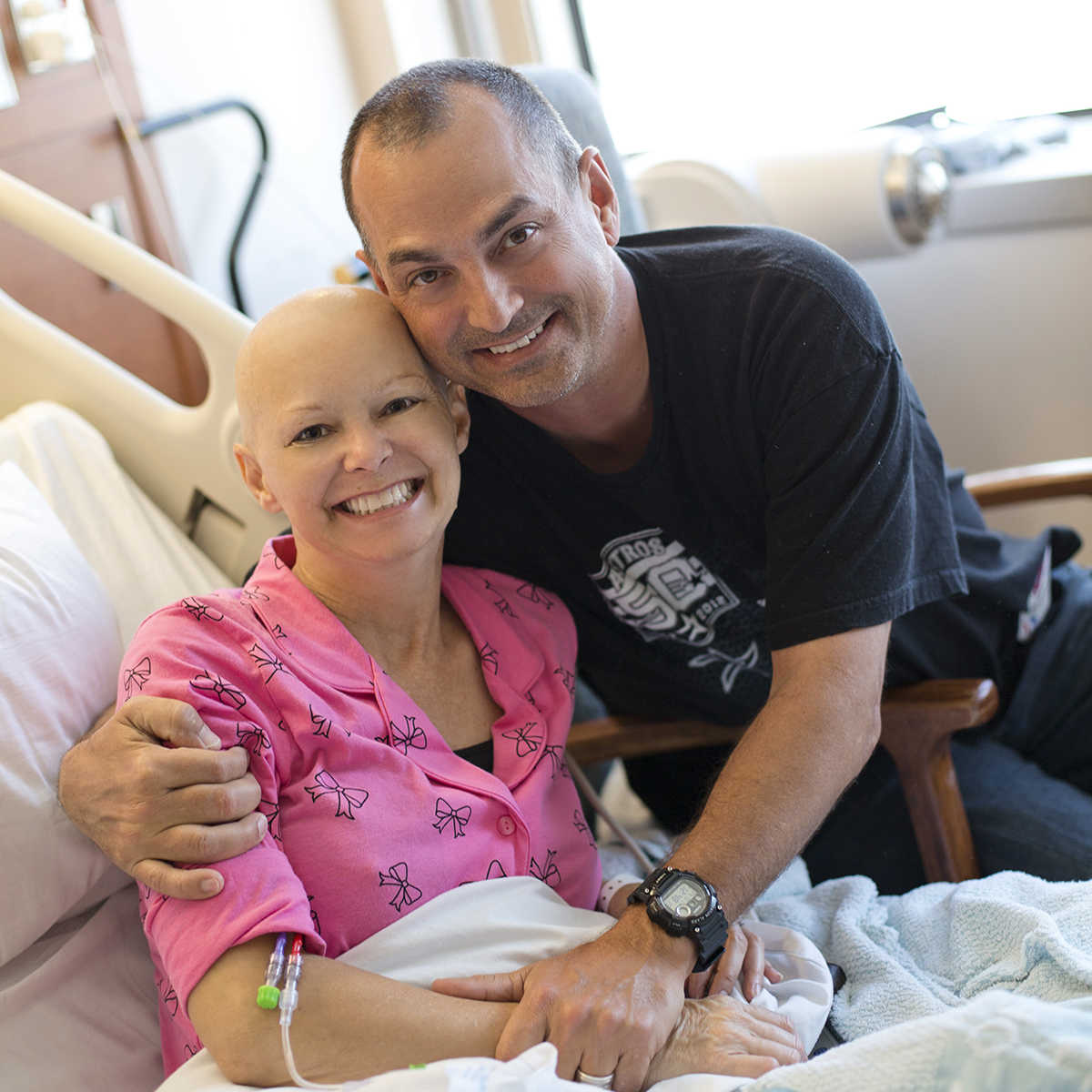 wife without hair sitting and smiling in hospital bed as husband has his arms around her smiling