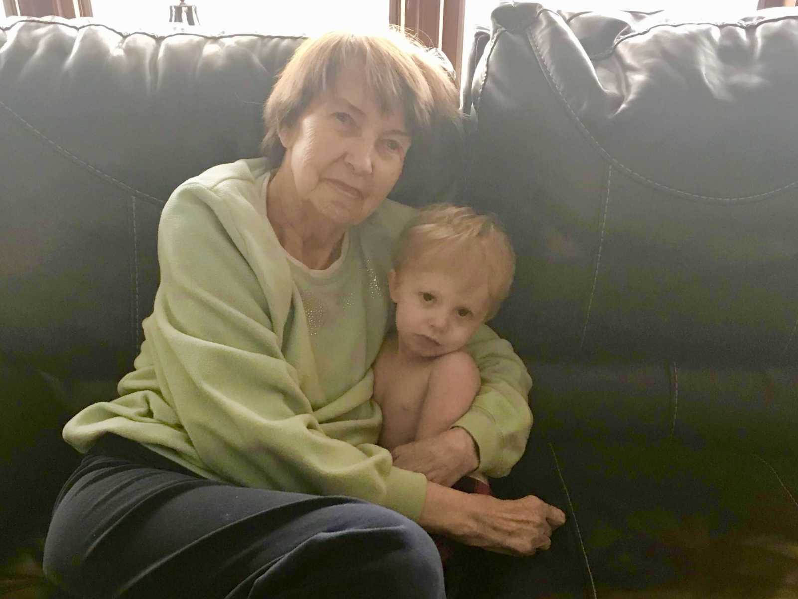 grandmother holds shirtless grandson in her arms as they sit on black leather couch