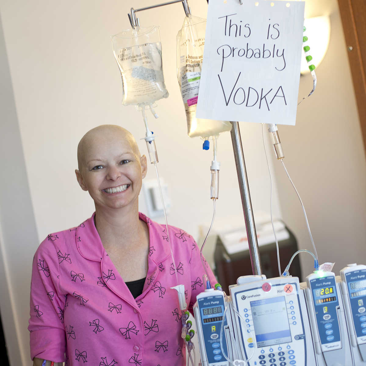 Smiling cancer patient standing next to an IV with sign that says, "this is probably vodka"