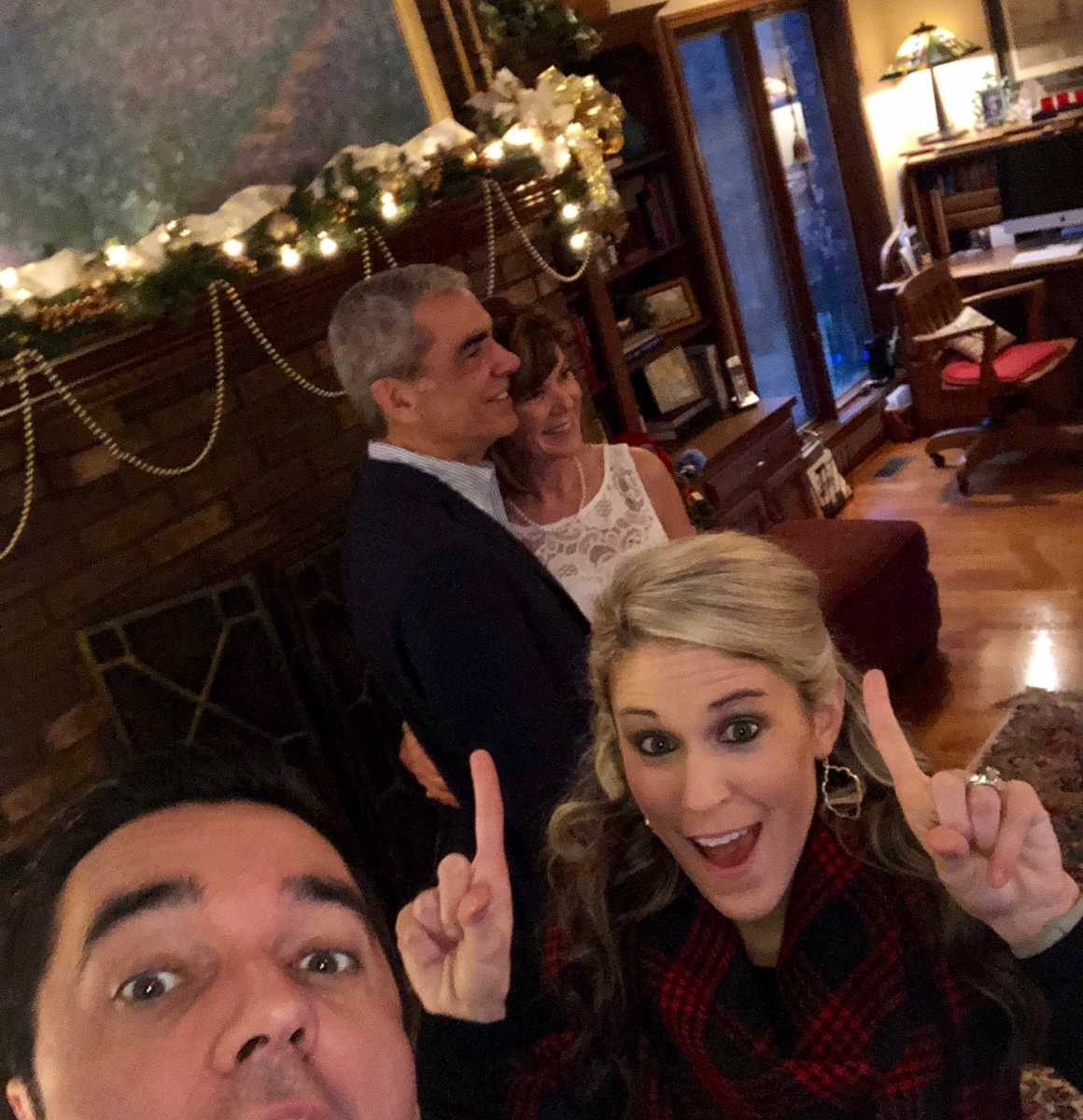 man takes selfie with woman holding up the number one with her fingers and married couple in front of chimney in background