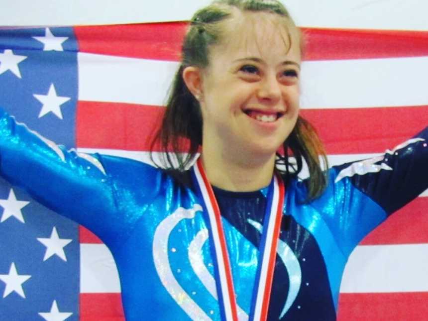 down syndrome girl in blue and white leotard smiling holding an american flag behind her and medal on her neck