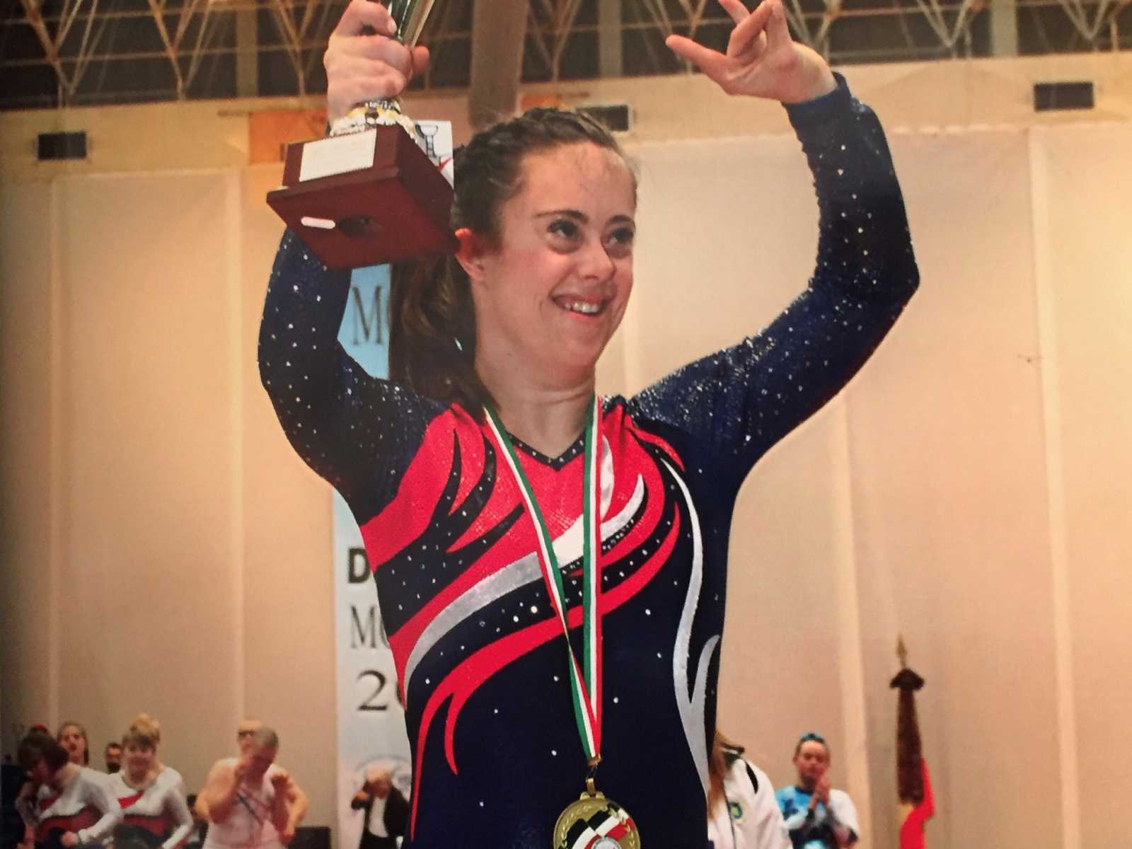 down syndrome girl in red white and blue leotard smiling holding a trophy in the air and a medal around her neck