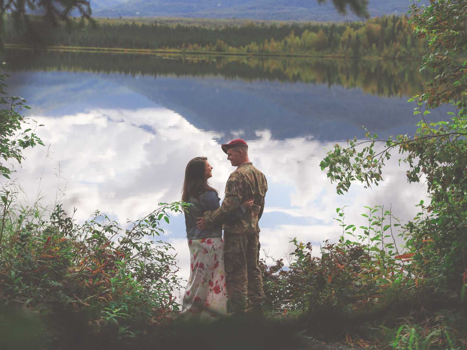 soldier and woman with arms wrapped around each other with water and trees in background