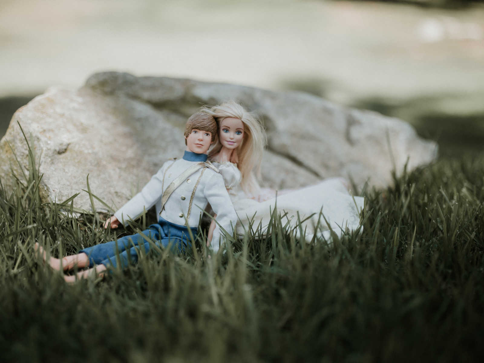 ken and barbie sitting in the grass leaning on each other in front of rock