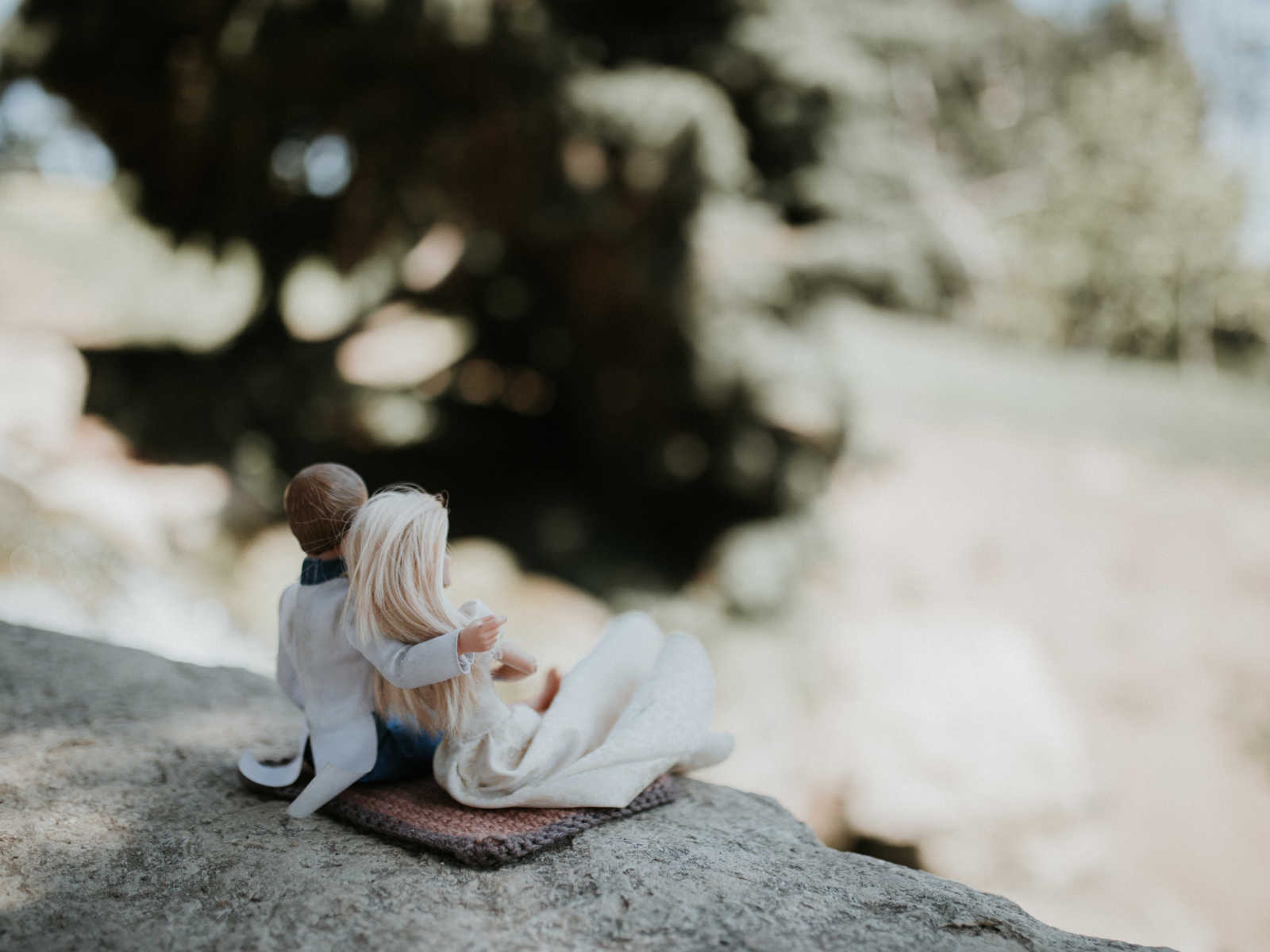 ken sits next to barbie on a rock with his arm wrapped around her while they look out into the distance
