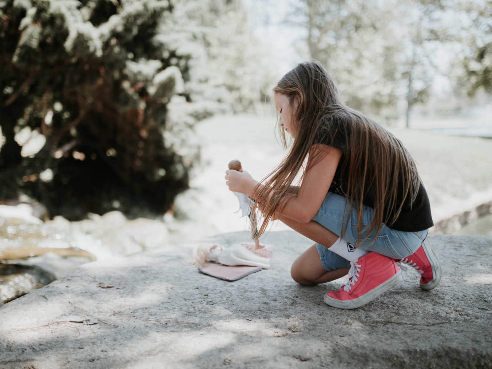 young girl kneels on large rock as she holds a ken doll and barbie is laying on the rock
