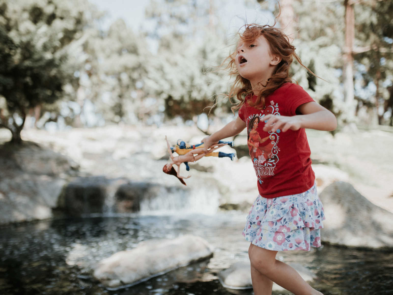 little girl with red curly hair holds two plastic dolls in front of large rocks and body of water