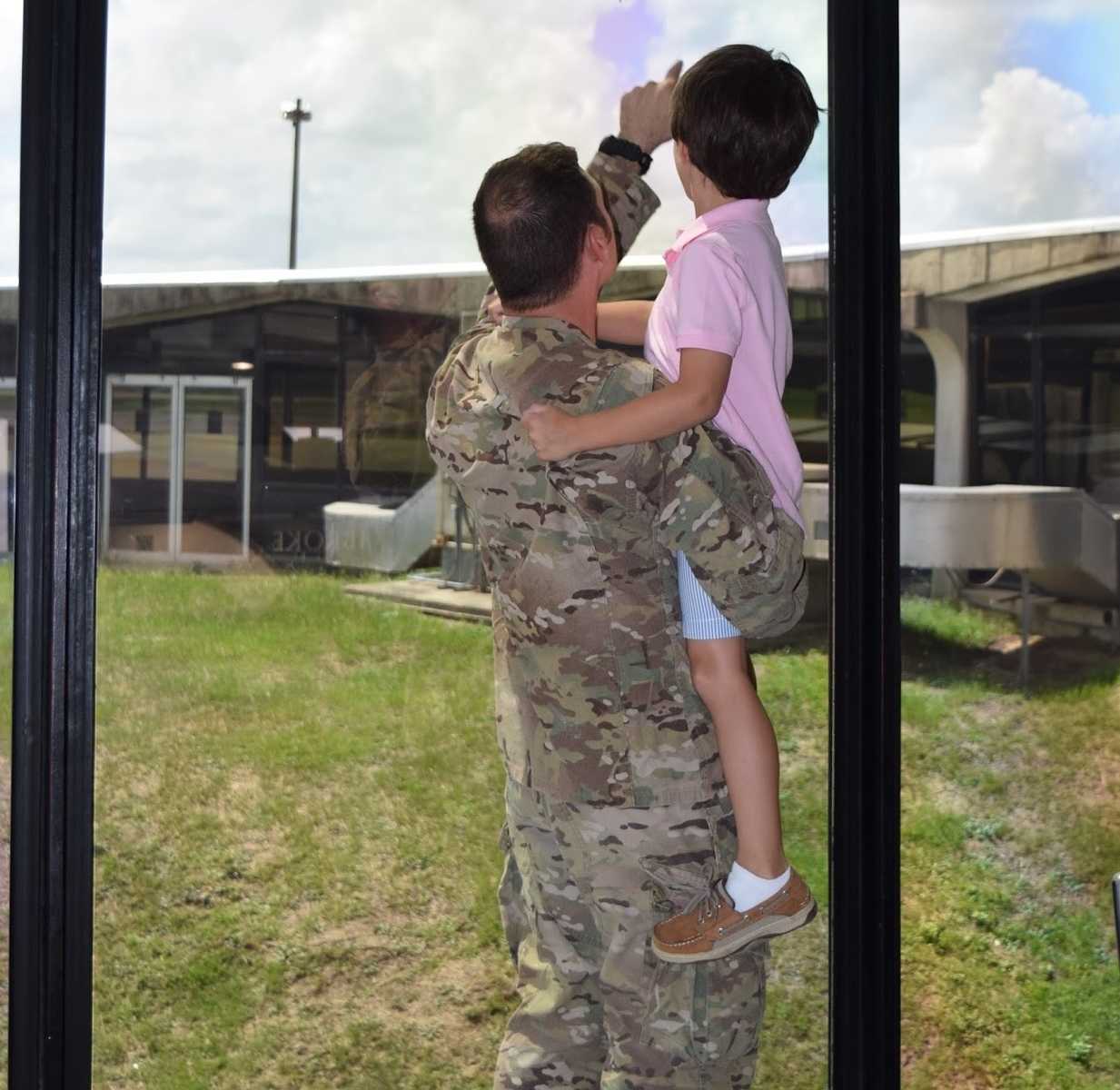 soldier in camo uniform holds toddler son while one arm points to something out the floor to ceiling window