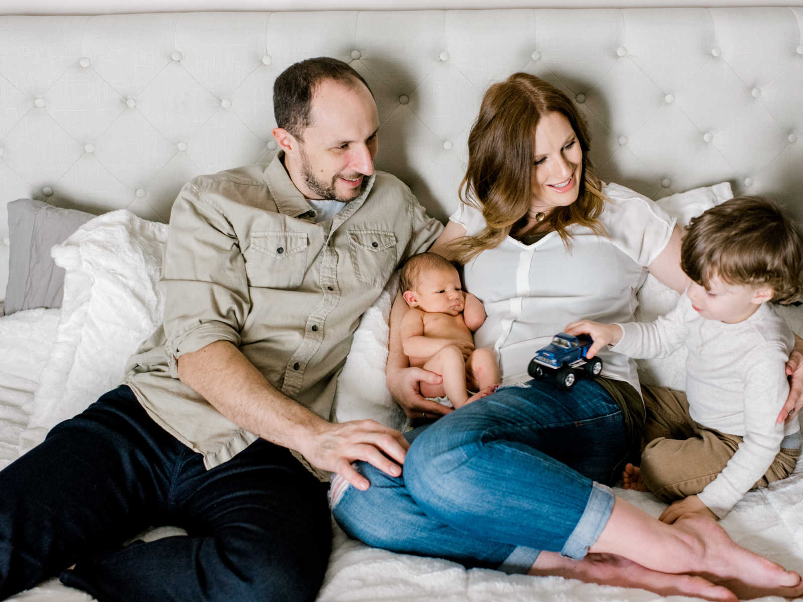 family sitting in bed with newborn in mothers arms while toddler plays with a toy car on moms leg