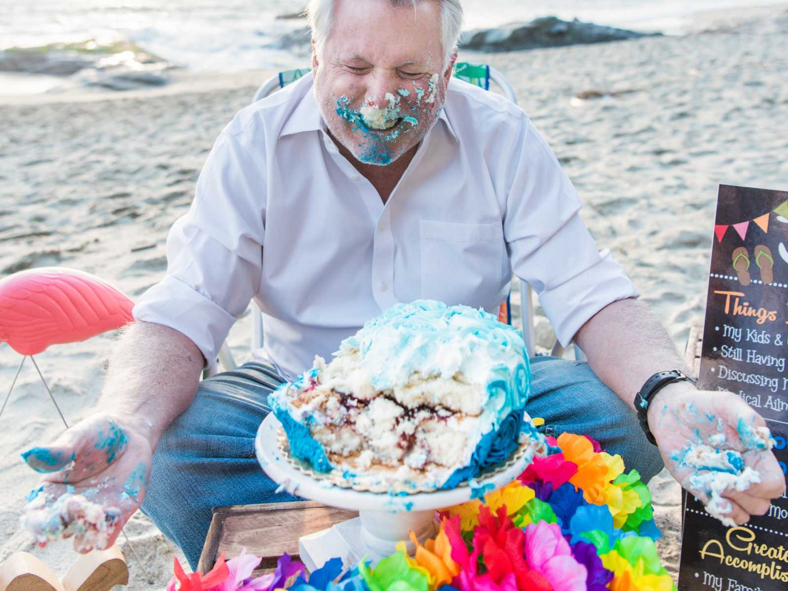 man smiles as he tears apart birthday cake that is now all over his face and hands on the beach