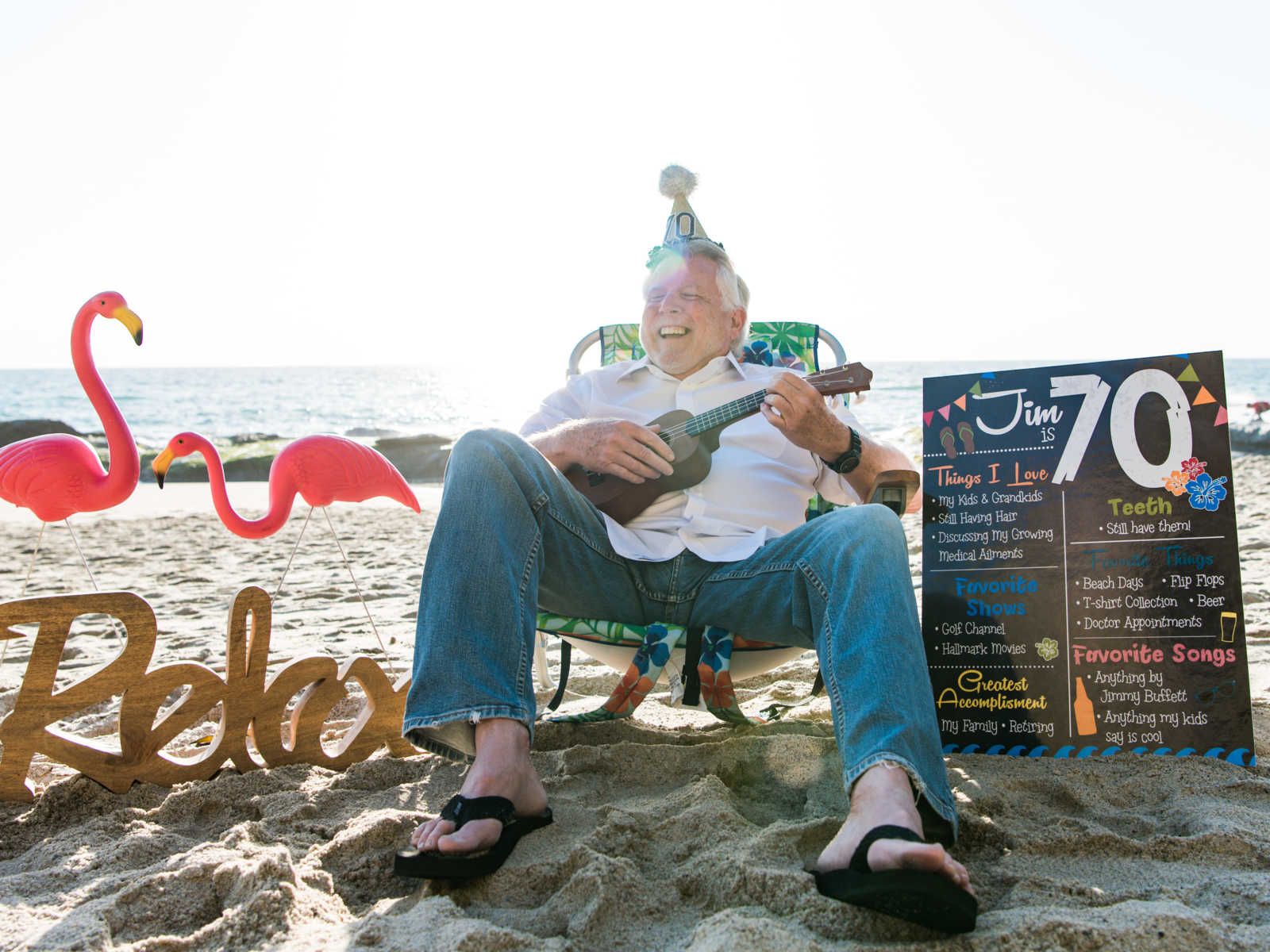 man with party hat sitting in beach chair smiling while playing the ukelele in the sand