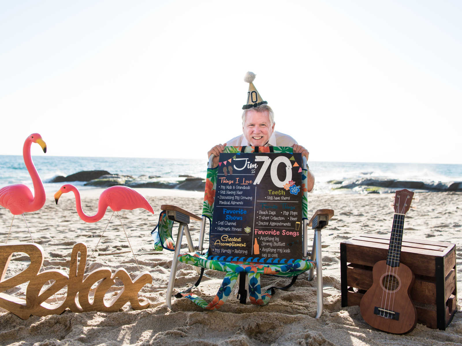 man kneeling behind beach chair in the sand with a party hat that has the number 70 on it with other beach themed props
