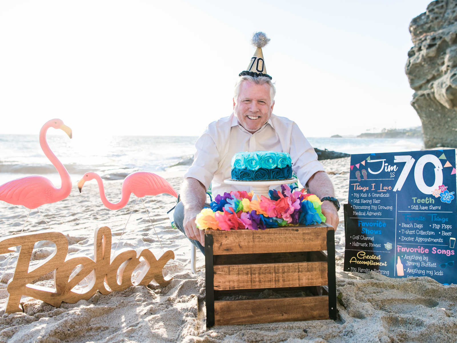 man smiles behind birthday cake covered in blue roses surrounded by colorful paper flowers