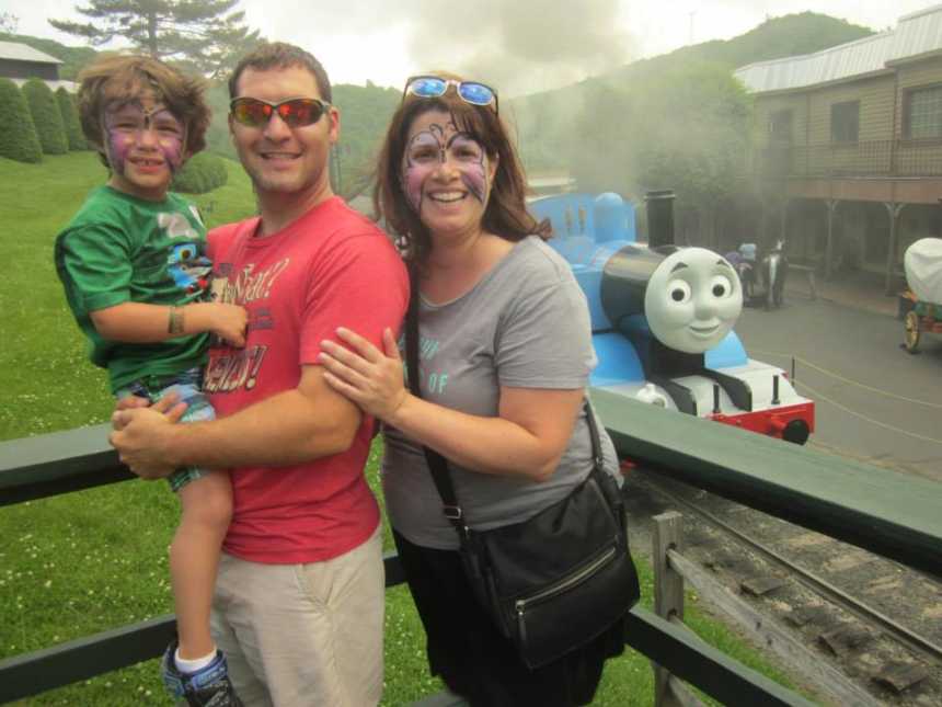 father stands holding son who has butterfly face paint next to wife with butterfly face paint in front of thomas the train 
