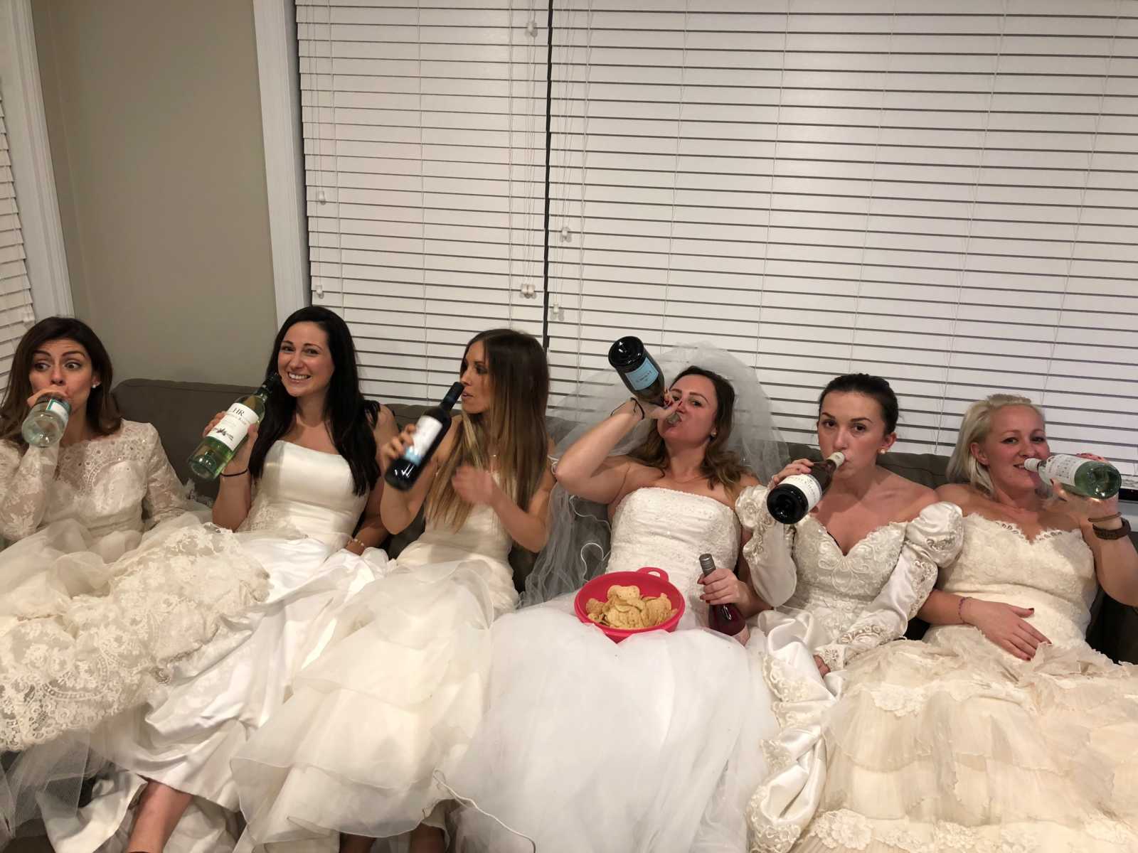 women sitting on a couch with wedding dresses on each holding their own bottle of wine