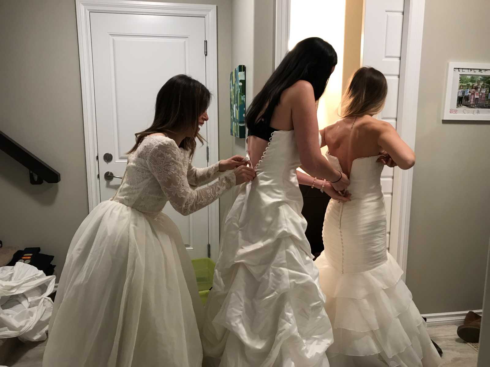 train of three women trying to zip and button up each others wedding gowns