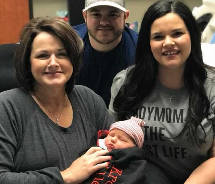 woman holds baby wrapped in a blanket in her arms with a man and woman sit next to her smiling