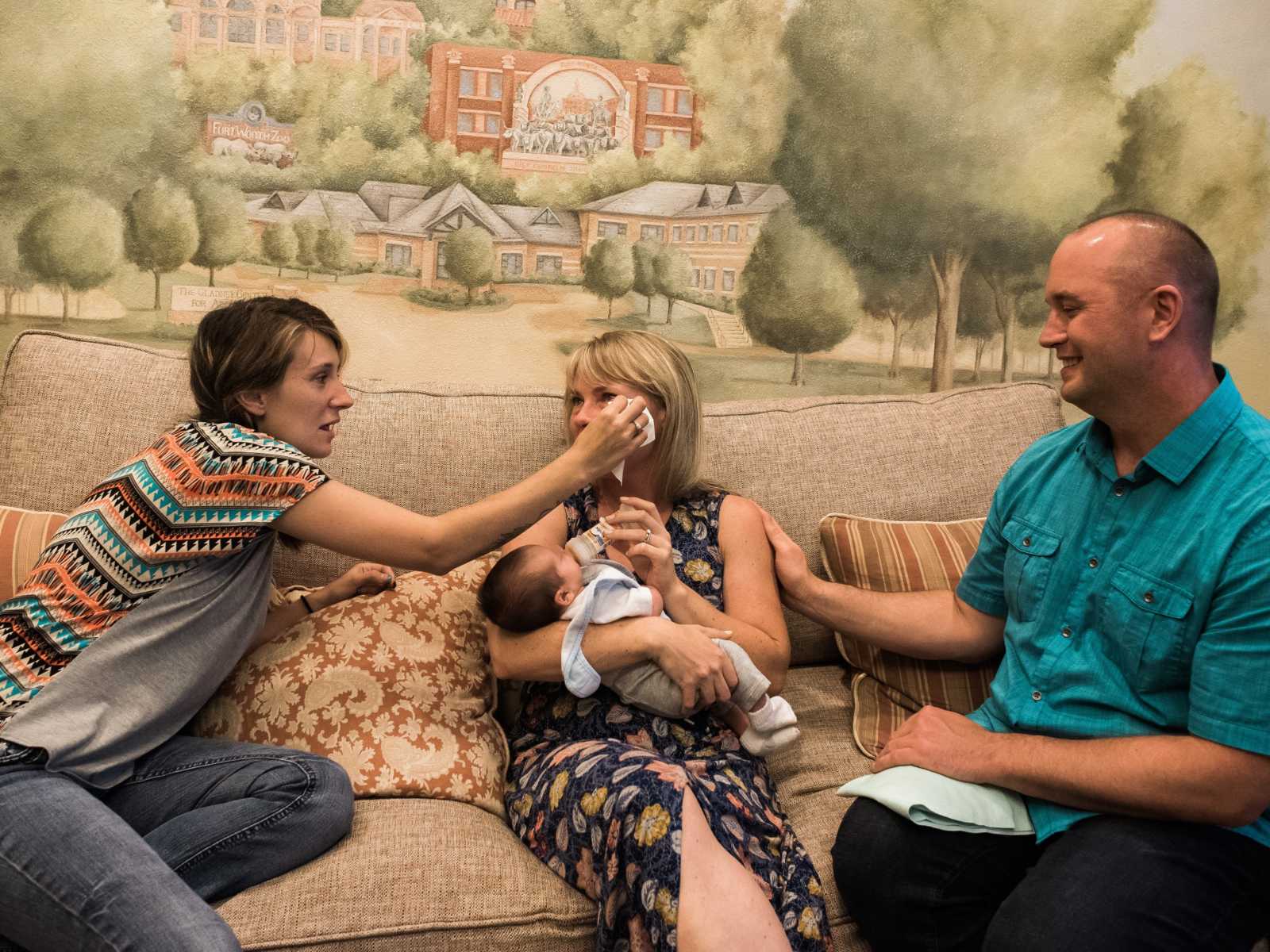 birth mother wipes tear away from adoptive mother holding son next to husband on couch 
