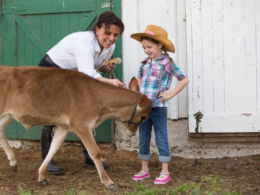 farm animal therapist smiles and leans over to pet calf next to young girl in cowboy hat with hand on her hips