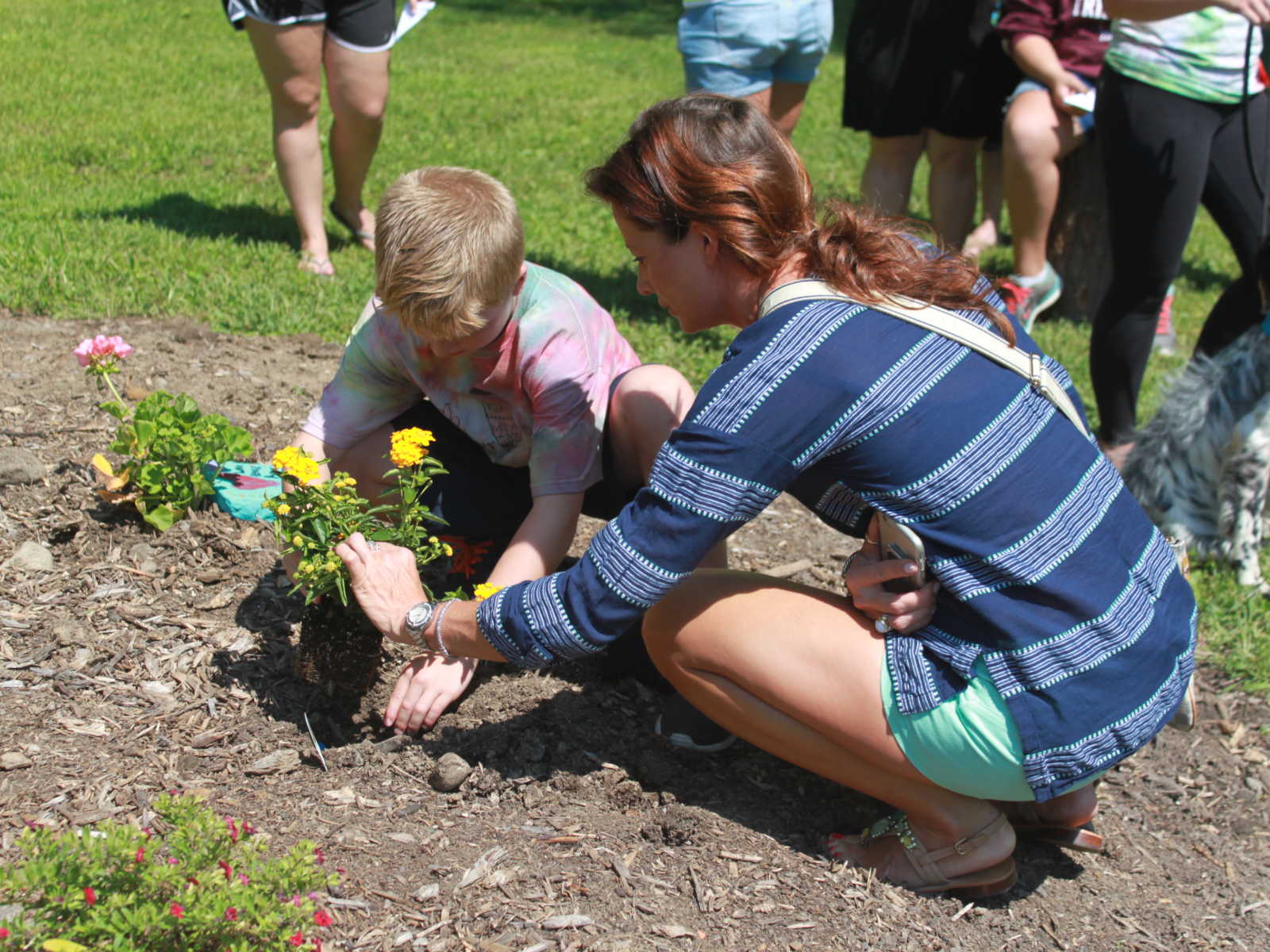 woman crouches down to help boy plant a yellow flower next to pink and red flower