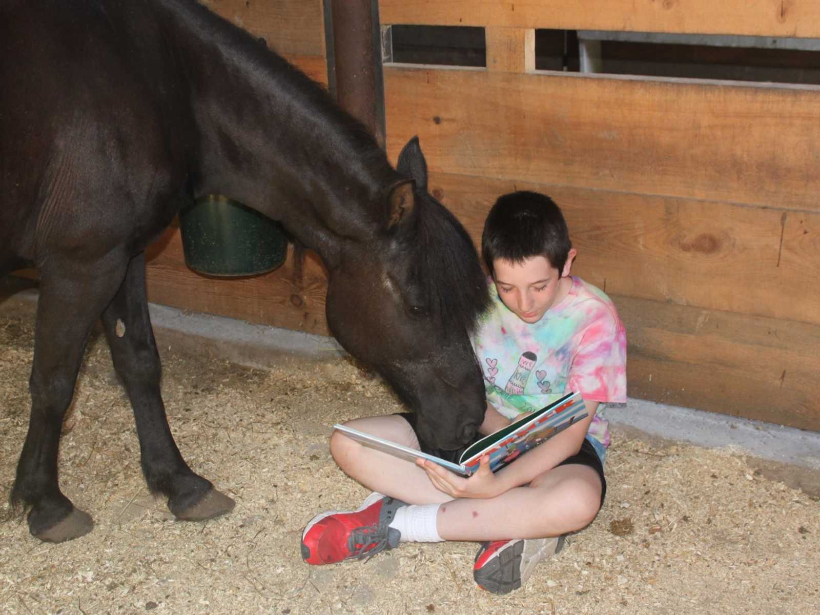 young boy sitting on ground of horse stall reading a book while horse leans over him with nose on book