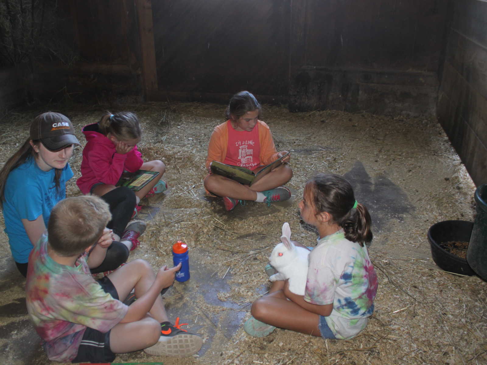 kids sitting in barn with teenager in a circle while on girl has white bunny in lap and two girls have books