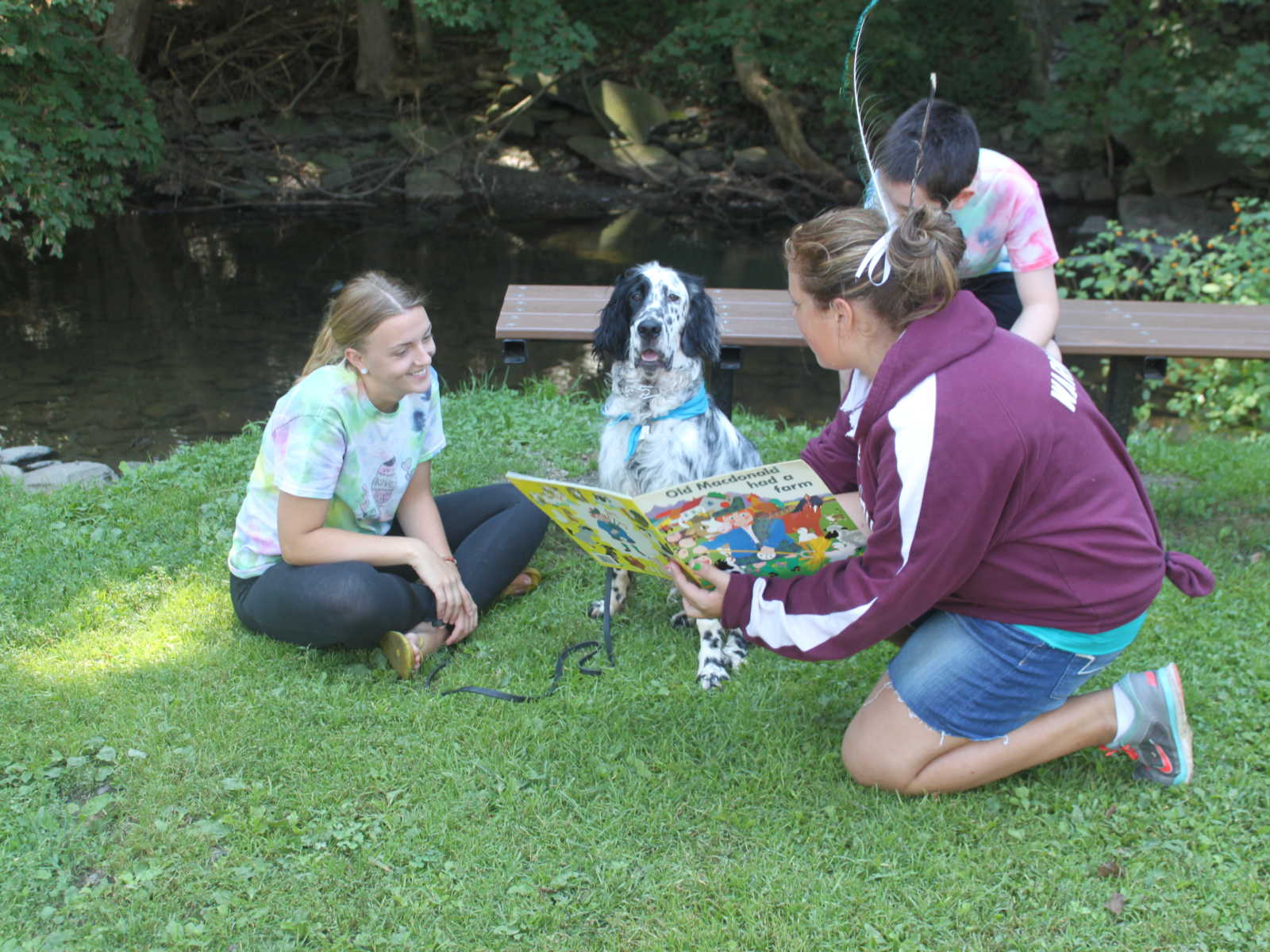 Teen girl kneels holding out a book to another teen girl who is sitting on the ground next to dog and boy on a bench